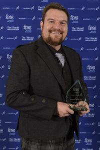 Gary, in a grey and black kilt, smart jacket and silver cravat, holding his award